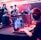 Several people are seated and focused on computer screens, wearing headsets, in an environment that appears to be a gaming tournament or esports event. The room is dimly lit with neon lighting, highlighting screens and creating an intense atmosphere.