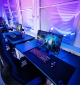 A row of modern gaming setups is arranged neatly along a window-lined wall. Each station includes a high-resolution monitor displaying vivid graphics, a mechanical keyboard with colored lighting, a gaming mouse, and an ergonomic chair. The room is illuminated with ambient blue and purple lights, creating a sleek and futuristic atmosphere.