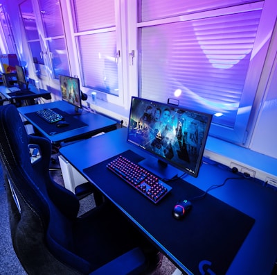 A row of modern gaming setups is arranged neatly along a window-lined wall. Each station includes a high-resolution monitor displaying vivid graphics, a mechanical keyboard with colored lighting, a gaming mouse, and an ergonomic chair. The room is illuminated with ambient blue and purple lights, creating a sleek and futuristic atmosphere.