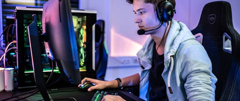 A young person is intensely focused on a computer screen while wearing a headset with a microphone. They are using a gaming keyboard and mouse, sitting in a gaming chair. The room has cool lighting, with a visible PC tower.