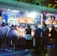 A group of people standing in front of an illuminated amusement arcade, engaging with various gaming machines. The environment is bustling with activity, and colorful lights create a vibrant atmosphere. The ceiling is decorated with large, playful images of dice and other game-related graphics, enhancing the lively mood.