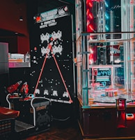 An arcade setting featuring a classic Space Invaders game setup and a modern gaming machine with seats. Bright neon lights illuminate the area, with scores displayed on digital screens. A bright and visually engaging environment full of electronic components.