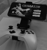 A mobile phone is mounted on a stand displaying a game titled 'Grimvalor' with options such as 'Continue', 'New Game', and 'Extras'. In front of the phone, there is a game controller placed on a table. The setup indicates gaming activity possibly using a smartphone as the screen.