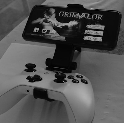 A mobile phone is mounted on a stand displaying a game titled 'Grimvalor' with options such as 'Continue', 'New Game', and 'Extras'. In front of the phone, there is a game controller placed on a table. The setup indicates gaming activity possibly using a smartphone as the screen.