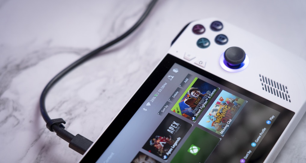 A handheld gaming console with a white body and colorful buttons features a screen displaying various game icons. The device is connected to a power source via a black cable and rests on a marble surface. The interface shows games like Apex and Street Fighter.