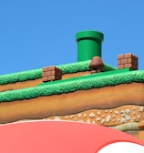 A colorful, stylized scene depicting a platform from a video game setting. The scene features a grass-topped surface with soil layers beneath, a green pipe, brick blocks, and a character resembling a mushroom. The sky is clear and blue, creating a vibrant backdrop.
