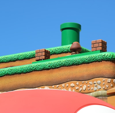 A colorful, stylized scene depicting a platform from a video game setting. The scene features a grass-topped surface with soil layers beneath, a green pipe, brick blocks, and a character resembling a mushroom. The sky is clear and blue, creating a vibrant backdrop.